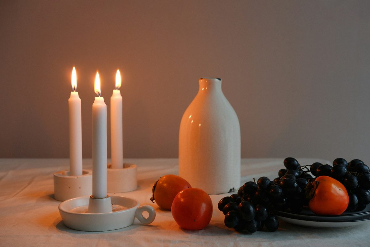 Trois bougies allumées, une bouteille en céramique blanche, des kakis et du raisin noir sur une nappe claire